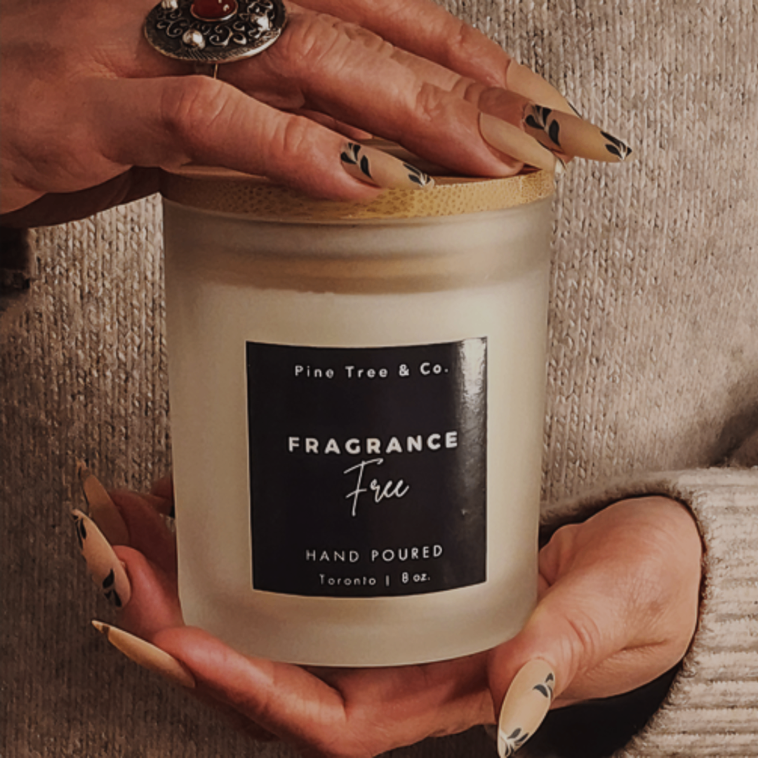 A person holding a fragrance-free soy wax candle with a label that reads 'Pine Tree & Co. Fragrance Free Hand Poured Toronto 8oz'.